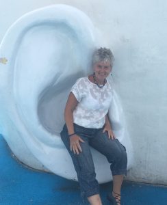 Foto van Maloush Köhler, een vrouw met kort grijs haar, die op een beeld van een heel groot wit oor zit