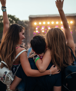 Drie jongeren vanaf de achterkant gezien. Een meisje, jongen en een meisje. Ze staan voor een podium en de meisjes hebben één arm in de lucht.