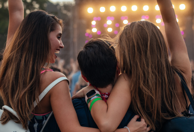 Drie jongeren vanaf de achterkant gezien. Een meisje, jongen en een meisje. Ze staan voor een podium en de meisjes hebben één arm in de lucht.