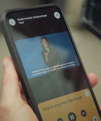 Mobiel in iemands hand waarop je ziet hoe een uitleg in gebarentaal wordt gegeven en waarop ondertiteling te lezen is.