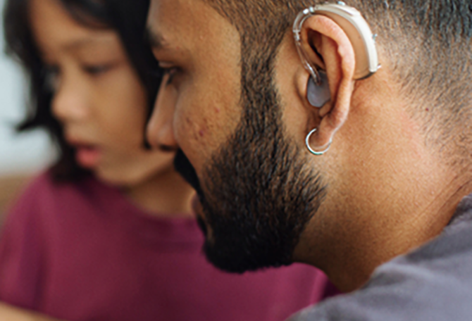 Close-up van een man vanaf de zijkant. Hij draagt een hoortoestel. Op de achtergrond zit een kind.