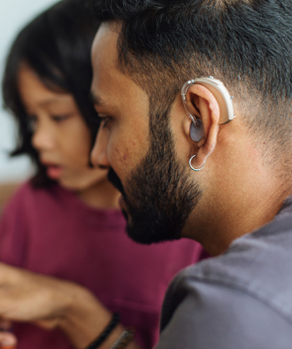 Close-up van een man vanaf de zijkant. Hij draagt een hoortoestel. Op de achtergrond zit een kind.